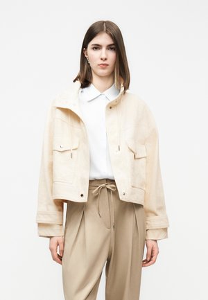 Ung person med glat brunt hår iført en lys beige kort jakke, hvid skjorte med krave og beige bukser med høj talje og snøre.