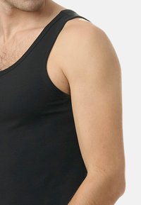 Manlig överkropp i en svart ärmlös tanktop, som visar axel, överarm och del av bröstet mot en ljus bakgrund.