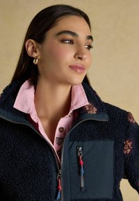 Marineblauwe fleecejas met bloemaccenten, zwart paneel en zakken met rits, gedragen over een roze overhemd met kraag. Soepele textuur en praktisch ontwerp.