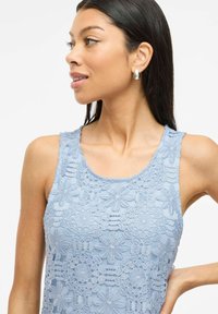 Woman wearing light blue sleeveless top with floral lace pattern, silver teardrop earrings, looking to the left with hand on hip.