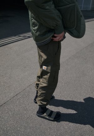 Person trägt olivfarbene Cargohose, schwarze Socken und schwarze Sandalen und steht bei Tageslicht mit den Händen nahe den Taschen auf Asphalt.