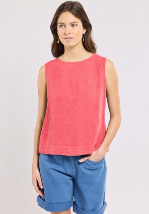 Femme portant un haut corail sans manches et un short bleu retroussé, debout avec une main dans la poche sur un fond uni.
