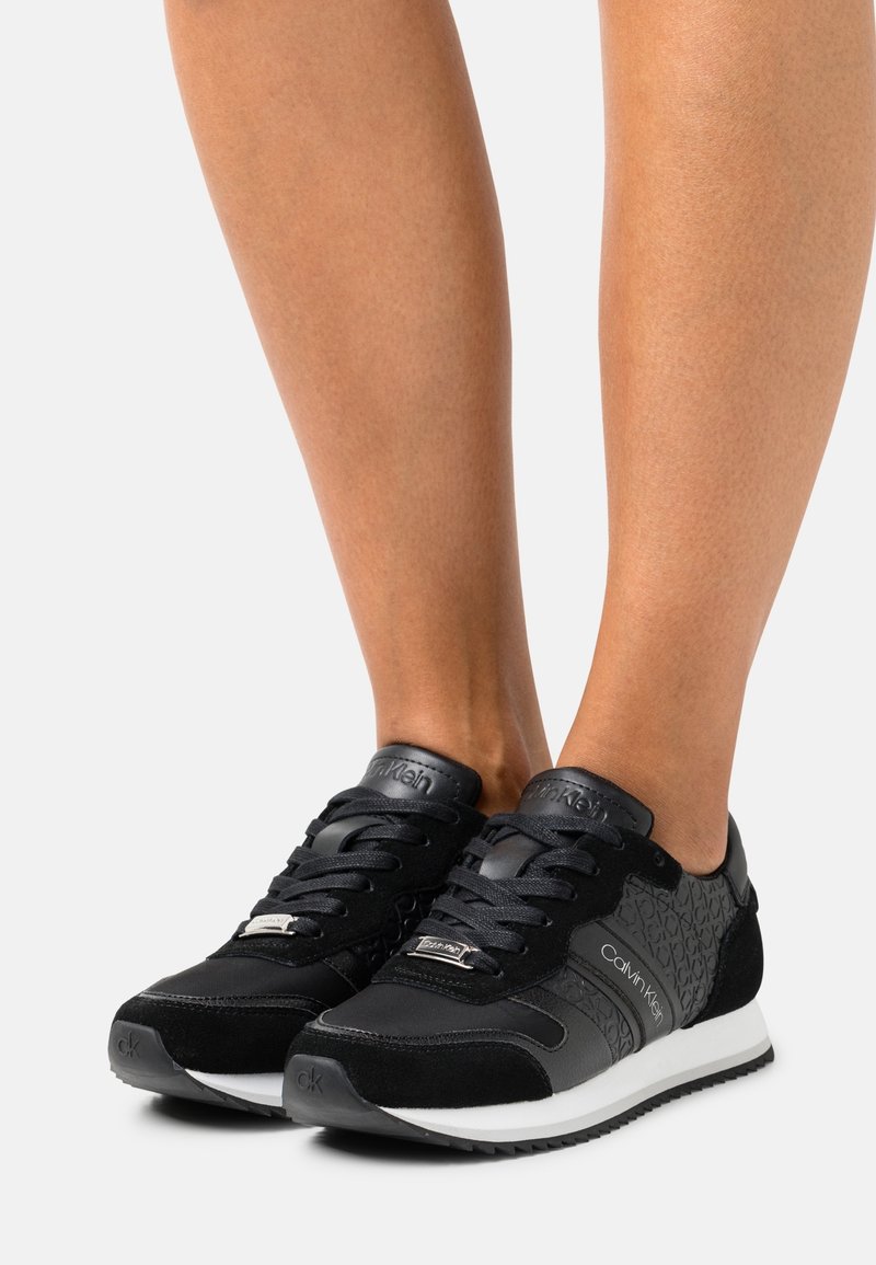 Baskets noires Calvin Klein avec lacets noirs et semelles blanches portées pieds nus sur un fond neutre.