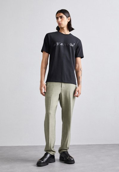 Black cotton t-shirt with a reflective graphic, paired with light green tailored trousers and black lace-up shoes.
