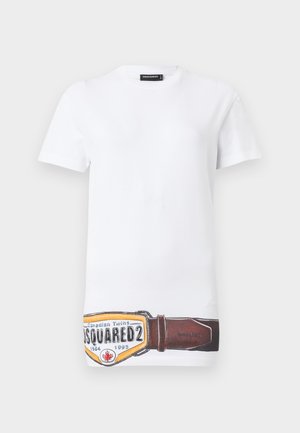 Γυναίκα που φοράει λευκό πουκάμισο με κοντά μανίκια, κόκκινο παντελόνι με φαρδιά μπατζάκια και floral εφαρμογή σε έναν μηρό, μαύρα σανδάλια και ζώνη με σχέδιο.