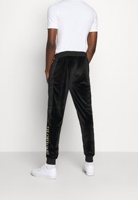 Eine Person steht mit dem Rücken zur Kamera, trägt ein weißes T-Shirt und schwarze Samtjogginghosen mit der Aufschrift "GLORIOUS" an der Seite, kombiniert mit weißen Turnschuhen.