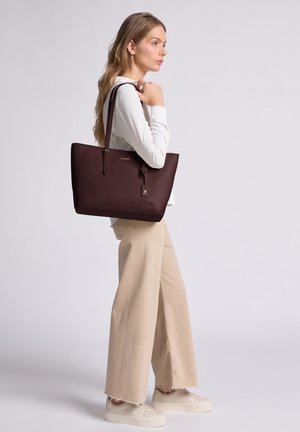 Woman with long hair wearing white top and beige wide-leg pants, carrying a large dark brown leather tote bag on shoulder.