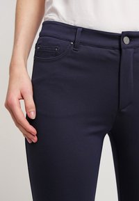 Marinblå stretchbyxor med en figursydd design, framfickor och en synlig knappstängning i midjan. Slät textur och skräddarsydd form.