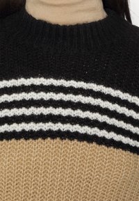 Schwarzer, weißer und beiger gestrickter Pullover mit horizontalen Streifen, getragen von einer Person in einem beigen Rollkragenpullover darunter.