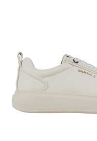 Sneaker bianco con tomaia in pelle liscia, punta tonda e suola in gomma. Caratterizzato da una marchiatura dorata, occhielli e una linguetta sul tallone.