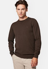 Brązowy sweter dzianinowy z okrągłym dekoltem, z długimi rękawami i ściągaczami. Posiada małe logo wyszyte na klatce piersiowej. Gładka faktura.