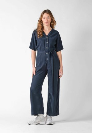 Young woman stands wearing navy blue buttoned jumpsuit with waist tie and white sneakers against plain light background.