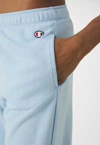 Světle modré tepláky s elastickým pasem a boční kapsou, s malým vyšitým logem v červené a černé barvě blízko pasu.