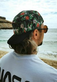 Hombre con gorra floral y gafas de sol mira hacia el mar en una playa de arena junto a una estructura de sombrilla de paja.