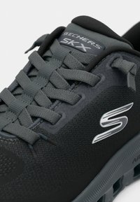 Scarpa sportiva nera con lacci grigi, tomaia testurizzata e suola ammortizzata. Presenta un logo argentato. Progettata per comfort e supporto.