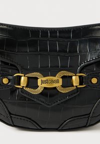 Crna torba od umjetne kože krokodila s zlatnim hardverom i "Just Cavalli" logotipom. Ima strukturni oblik i detaljne šavove.