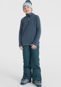 Fleecepullover i blått, med en fjärdedels dragkedja; ihop parat med teal-färgade snöbyxor och grå stövlar med svart hårdvara och blå accenter.