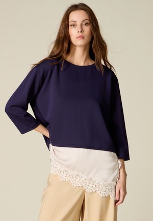 Donna con capelli castani lunghi indossa un top largo blu navy sopra una sottoveste in raso crema con bordi in pizzo e pantaloni beige, posando con una mano sul fianco.