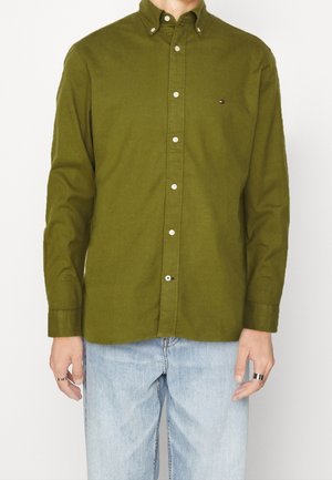 Homme portant une chemise boutonnée vert olive avec un petit logo sur la poitrine et un jean bleu clair, debout sur fond blanc.