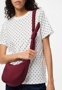 Borsa a tracolla bordeaux dalla forma morbida e arrotondata con tracolla annodata; indossata sopra una camicia bianca a pois, evidenziando un design casual.