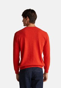 Mężczyzna z krótkimi ciemnymi włosami, ubrany w jasnoczerwoną bluzę i ciemnoniebieskie dżinsy, stojący tyłem do kamery na jednolitym tle.