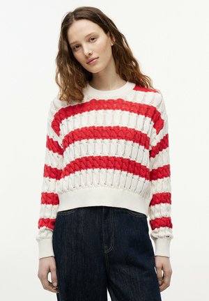 Jeune femme aux cheveux bruns ondulés portant un pull blanc à rayures horizontales rouges et un jean bleu foncé, debout devant un fond uni.