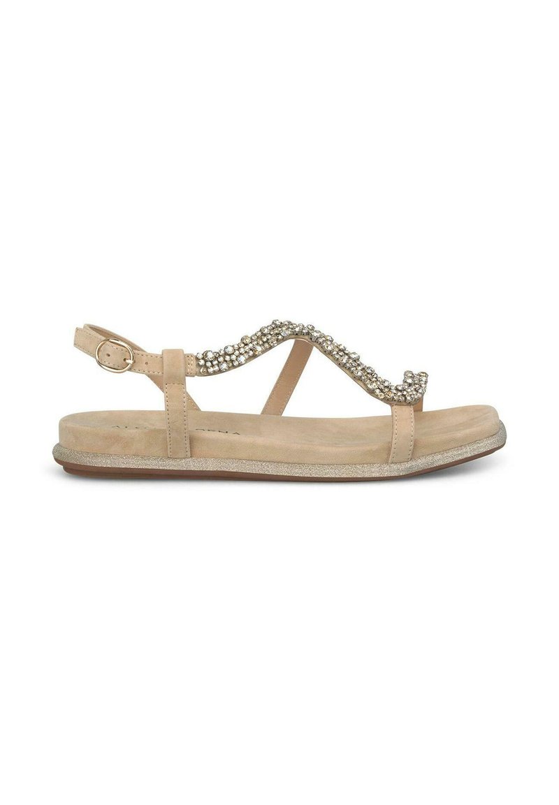 Sandalia beige con suela plana, que presenta una onda decorativa de piedras preciosas transparentes en la correa y una hebilla ajustable en el tobillo.