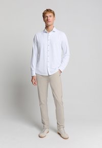 Witte langslavige overhemd met knopen, van gladde stof, gecombineerd met beige broek en lichte sneakers, staand tegen een neutrale achtergrond.