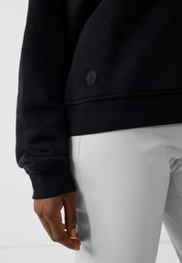 Felpa nera con tessuto testurizzato e polsini a coste, dotata di una toppa rotonda nera con logo. Abbinata a pantaloni grigio chiaro.