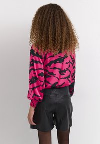 Bluzka z długim rękawem w jaskrawym różowym satynie z czarnym abstrakcyjnym wzorem, połączona z czarnymi skórzanymi szortami. Posiada marszczone mankiety i gładką fakturę.