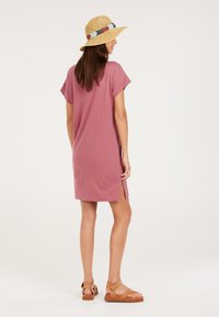 Rosa Baumwollkleid mit kurzen Ärmeln, Seitenschlitzen und lockerer Passform. Kombiniert mit einem beigefarbenen geflochtenen Hut und braunen Sandalen, vor einem neutralen Hintergrund.