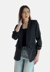 Blazer nero con colletto a risvolto, due tasche frontali e maniche arrotolate, abbinato a jeans in denim a vita alta di colore azzurro chiaro.