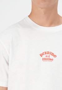 Witte katoenen t-shirt met rood grafisch tekst: "PERDIDO en el PARAÍSO" met kleinere tekst eronder, korte mouwen, ronde halslijn.