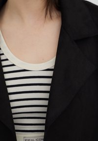 Personne portant une veste noire par-dessus un shirt à rayures horizontales noires et blanches avec un col rond, cou et haut de la poitrine visibles.