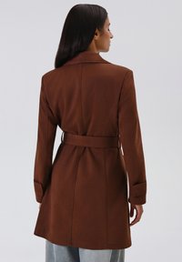 Cappotto marrone con cintura, dal design sartoriale, maniche lunghe e texture liscia. Presenta una fessura sul retro e una silhouette snella.