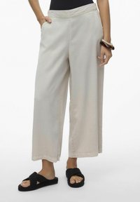 Pantaloni a gamba larga di un leggero beige, con tasche laterali, in tessuto morbido e vestibilità rilassata, abbinati a sandali neri intrecciati indossati a piedi nudi.