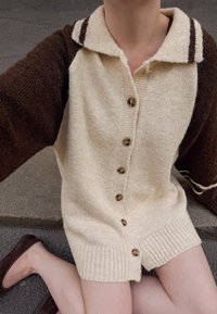 Gaišbrūns un brūns adīts kardigāns ar apkakli, kas aprīkots ar sešām pogām, ribotu apakšmalu un kontrastējošas krāsas piedurknēm. Mīksta tekstūra, ikdienišķs dizains.