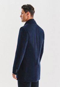 Granatowy wełniany płaszcz o dopasowanym kroju, z klapami i mankietami na trzy guziki. Miękka tekstura i czyste linie podkreślają elegancki design.