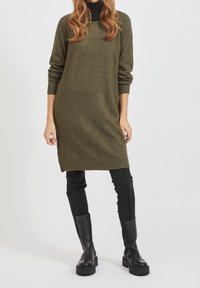 Abito lavorato a maglia verde oliva con maniche lunghe, polsini e orlo a coste, abbinato a pantaloni neri aderenti e stivali neri fino al ginocchio.