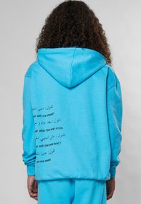 Sudadera color turquesa con capucha ajustable, que presenta texto en varios idiomas en la parte de atrás. Tejido suave y corte relajado con puños acanalados.