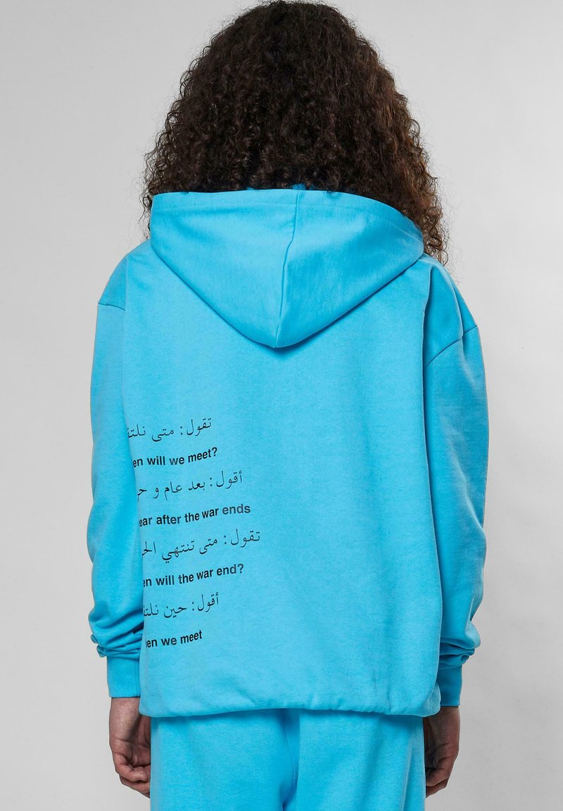 Sudadera color turquesa con capucha ajustable, que presenta texto en varios idiomas en la parte de atrás. Tejido suave y corte relajado con puños acanalados.