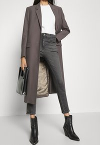 Un largo blazer gris sobre una blusa blanca, combinado con unos jeans negros de cintura alta. Unas botas de tacón negras hasta el tobillo y un bolso negro completan el look.