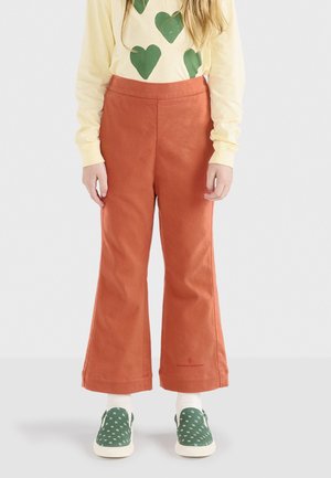 Pantalon évasé couleur rouille avec une texture lisse, associé à une chemise à manches longues jaune ornée de motifs en forme de cœur verts et des chaussures à pois verts.