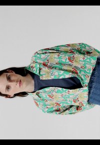 Jeune personne aux cheveux longs et foncés portant une veste à motif floral par-dessus une chemise sombre et un jean bleu, sur un fond uni.
