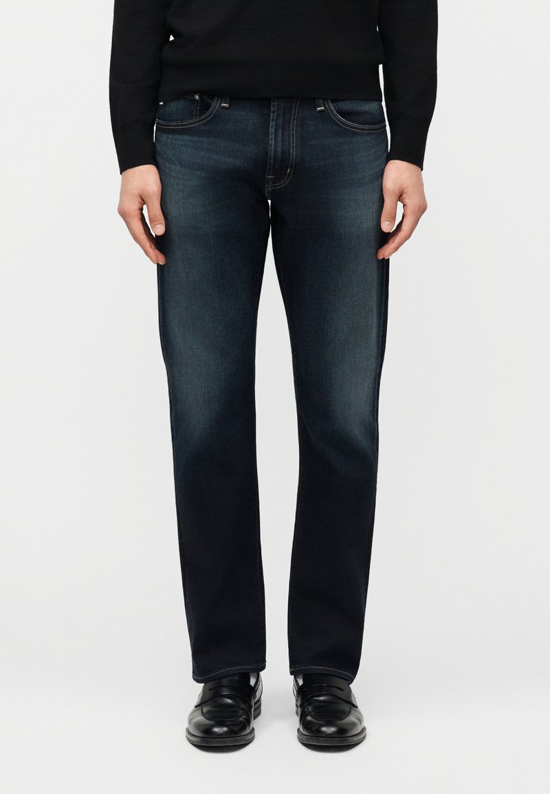 Άνδρας που φοράει σκούρα μπλε τζιν με ίσια γραμμή, μαύρο πουλόβερ και μαύρα δερμάτινα παπούτσια, στέκεται μπροστά σε απλό λευκό φόντο.