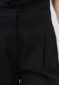 Personne portant un pantalon noir et une chemise noire, vue de la taille à la partie supérieure de la cuisse, avec un devant plat et des passants de ceinture visibles.