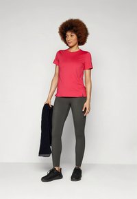 Růžové sportovní tričko s krátkým rukávem v kombinaci s tmavě zelenými legínami. Modelka má na nohou černé tenisky a drží tmavě modrou bundu.