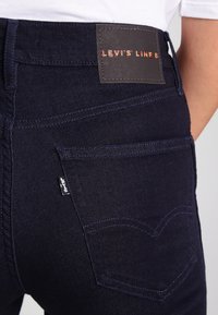 Jeans de mezclilla oscura con un parche que dice "LEVI'S LINE 8" y una pequeña etiqueta de marca. Detalle distintivo de costura en el bolsillo trasero visible.