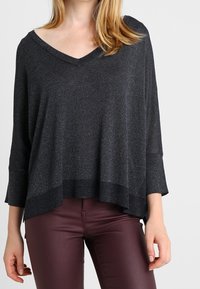 Czarny, luźny sweter z dekoltem w serek, rękawami do połowy przedramienia i żebrowanym dołem. Tkanina ma subtelny połysk. Połączony z bordowymi spodniami.