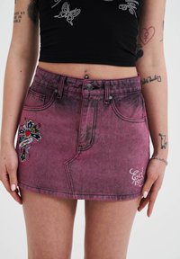 Korte roze denimrok met een vervaagde textuur, voorzien van een kleurrijk geborduurd kruisontwerp aan de voorkant en decoratieve stiksels.
